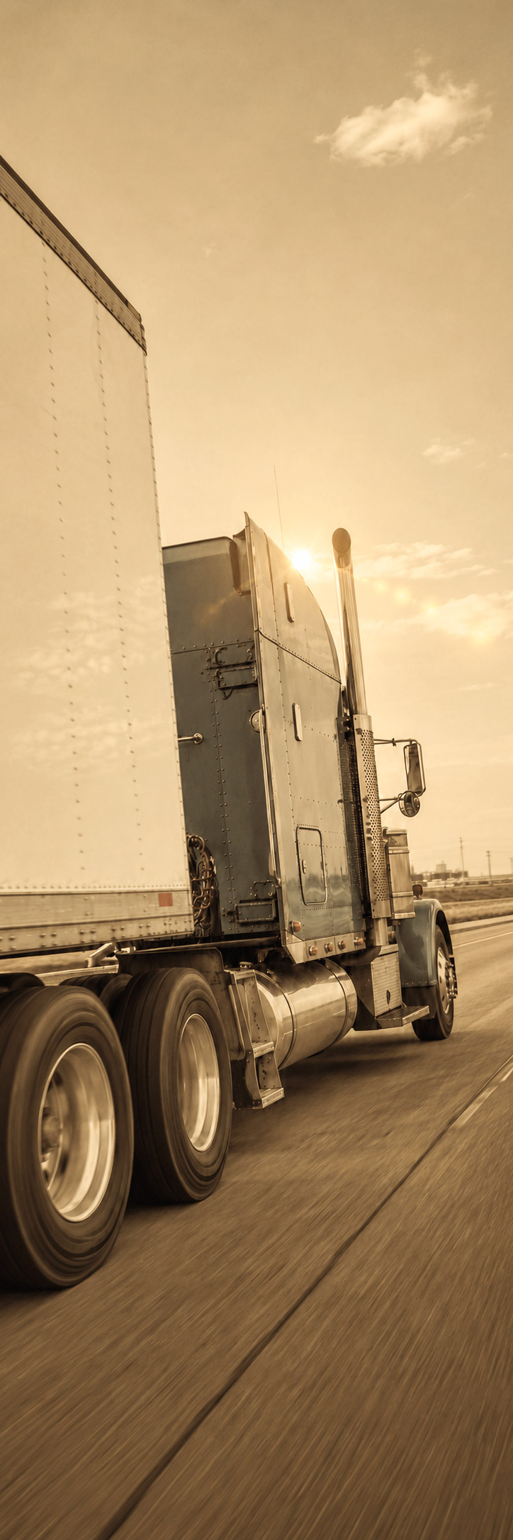 Rear three-quarter view of a semi-truck hauling a white trailer along a highway, with the low sun shining between the cab and exhaust stack.